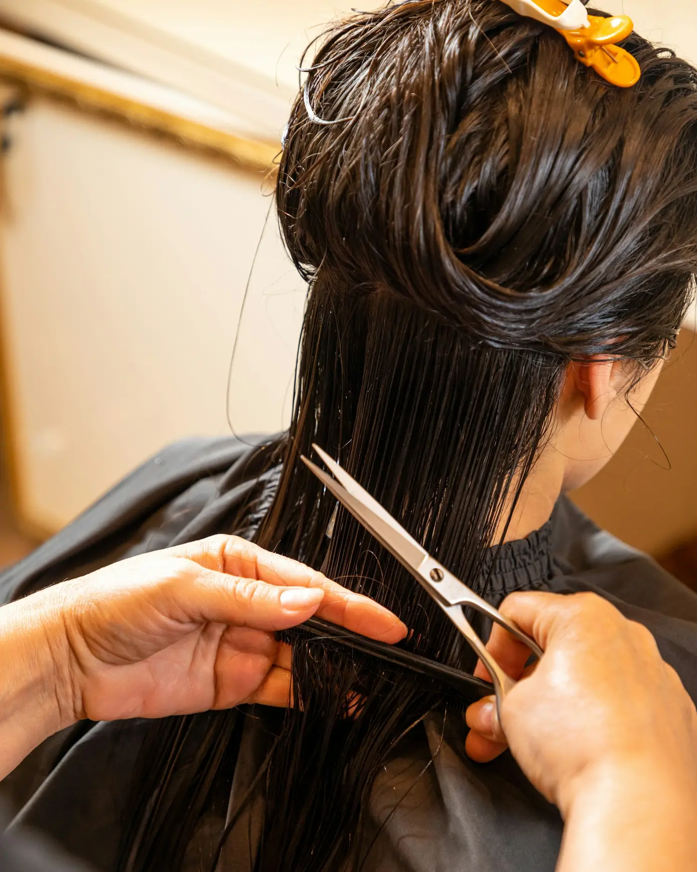 Mãos cortando uma mecha de cabelo escuro com tesoura.