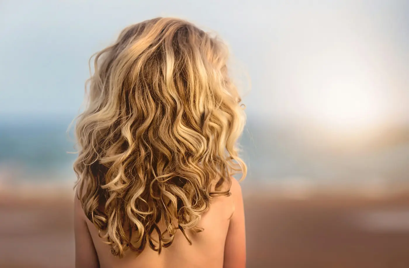 Cabelo loiro dourado e cacheado, visto de costas contra a luz.